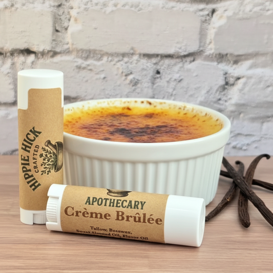 Two lip balm tubes labeled 'Hippie hick' Apothecary' next to a bowl of crème brélé on a wooden surface with a brick wall background.