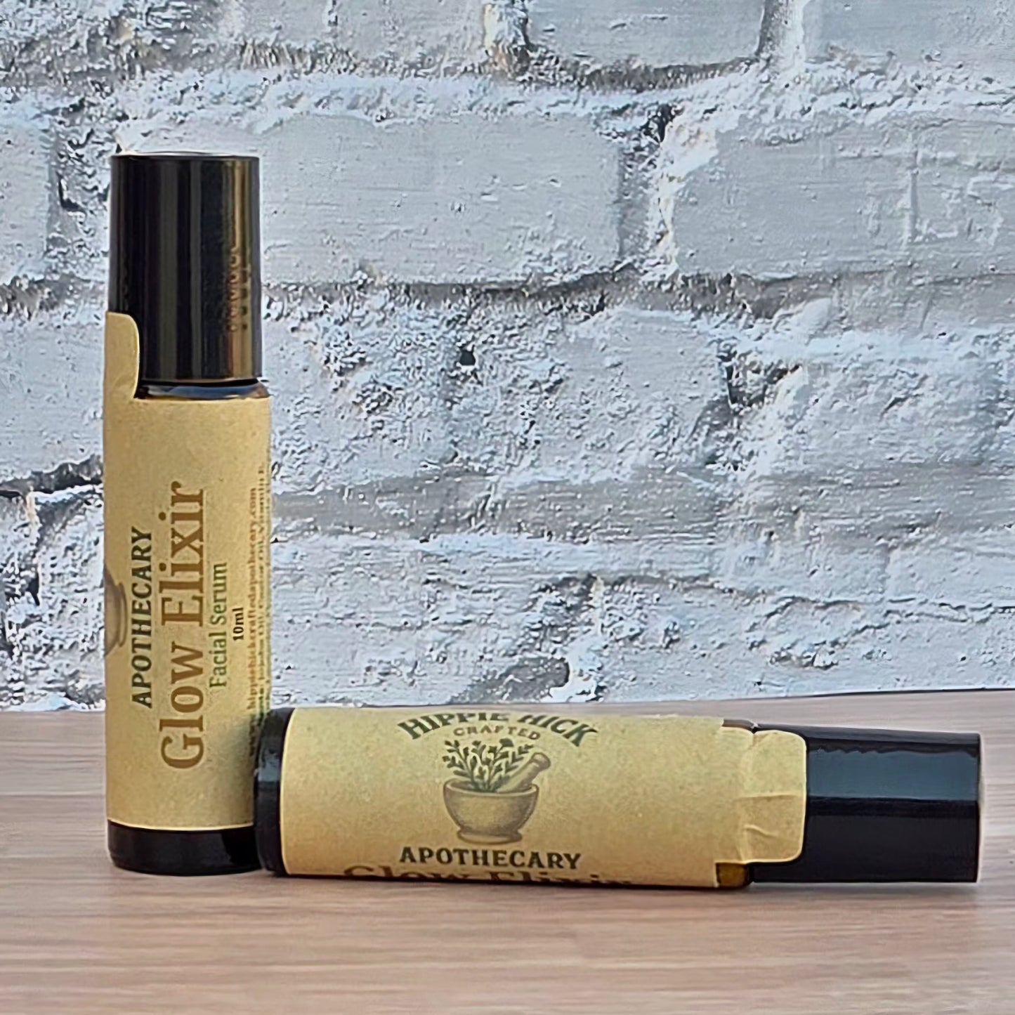 Two small cosmetic tubes labeled 'Apothecary Glow Elixir' with black caps and tan paper labels, placed on a wooden surface against a white painted brick wall.
