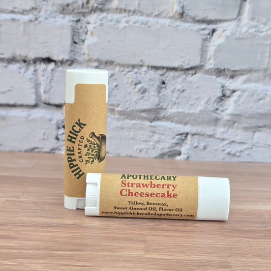 Two strawberry cheesecake tallow lip balm sticks with 'Hippie Hick' branding on a wooden surface with a brick wall background.