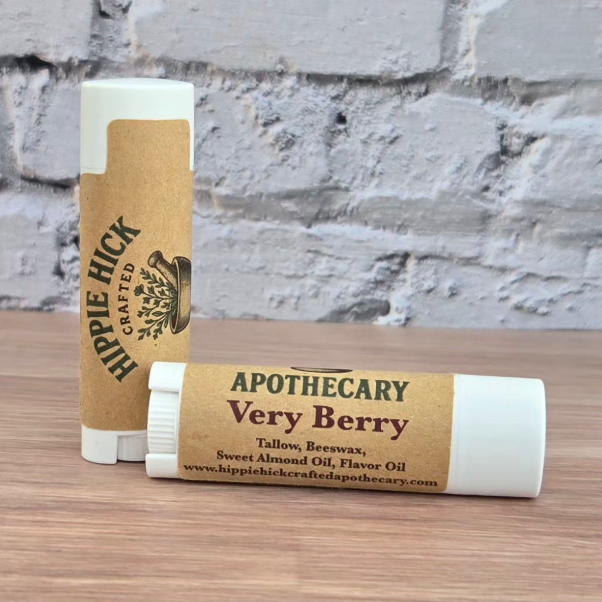 Two tallow lip balm sticks with brown paper wraps on a wooden surface against a brick wall background.